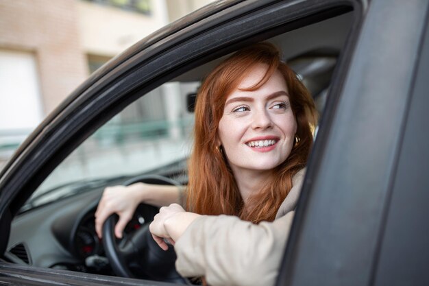 Mujer pelirroja alegre dentro del auto mirando hacia atrás desde el asiento del conductor mientras conduce durante el día