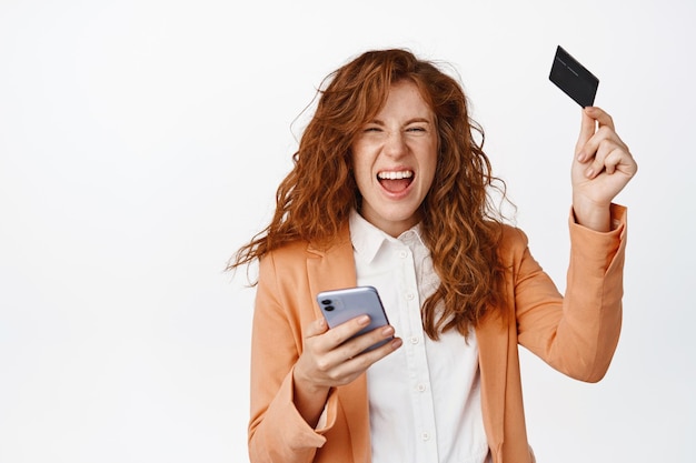 Mujer pelirroja alegre en traje que muestra tarjeta de crédito y teléfono inteligente gritando asombrada haciendo dinero en línea usando la aplicación móvil de pie sobre fondo blanco