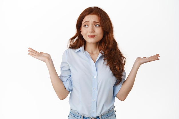 Mujer pelirroja ansiosa encogiéndose de hombros indecisa, mirando a un lado con una mirada confusa de pánico, no sabe nada, no puede entender, no tiene ni idea, de pie contra el fondo blanco