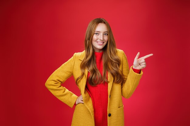 Mujer pelirroja atractiva elegante y segura de sí misma con pecas en abrigo amarillo al aire libre apuntando a la derecha con pistola de dedo y sonriendo con alegría como cámara mostrando una promoción genial sobre la pared roja.