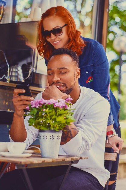 Mujer pelirroja atractiva y hombre de sonrisas negras usando un teléfono inteligente en la mesa en un café en una calle.