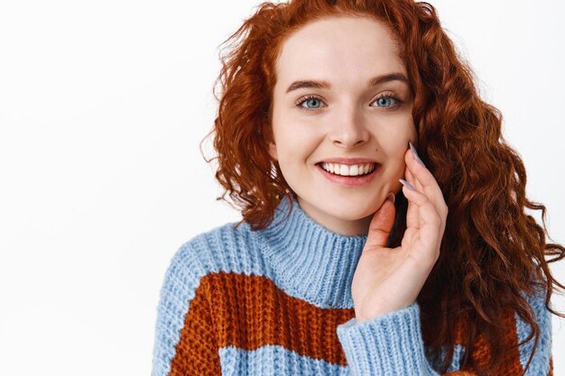 Mujer pelirroja con cabello rizado y rostro hermoso, dientes blancos sonrientes y tocando la piel facial suave y limpia, publicidad de productos de rutina para el cuidado de la piel o champú para el cuidado del cabello en blanco