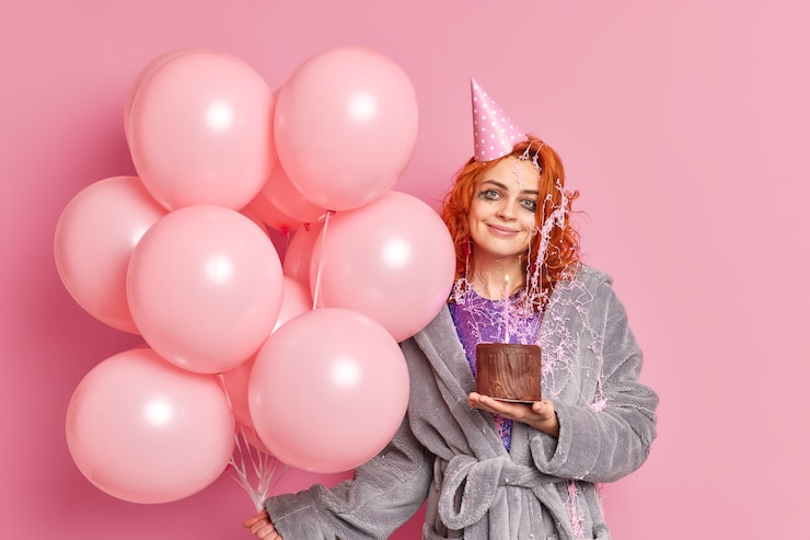Foto gratuito la mujer pelirroja complacida sostiene el pastel de chocolate celebra el aniversario se divierte en la fiesta vestida con ropa casual sostiene un montón de globos de aire inflados de color rosa tiene una expresión alegre