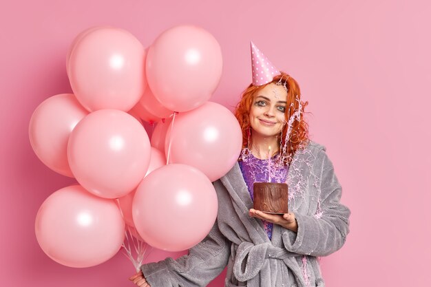 La mujer pelirroja complacida sostiene el pastel de chocolate celebra el aniversario se divierte en la fiesta vestida con ropa casual sostiene un montón de globos de aire inflados de color rosa tiene una expresión alegre