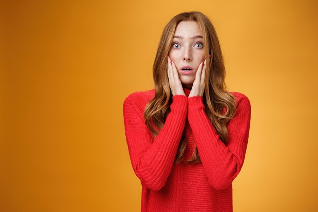 Mujer pelirroja conmocionada preocupada reaccionando a noticias problemáticas impresionantes sosteniendo las palmas en las mejillas, boca abierta y levantando las cejas asombrada, posando preocupada contra el fondo naranja