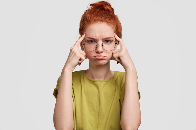 La mujer pelirroja decepcionada mantiene los dedos índice en las sienes, tiene la cara hosca, frunce el labio inferior, piensa en algo vestido de manera informal