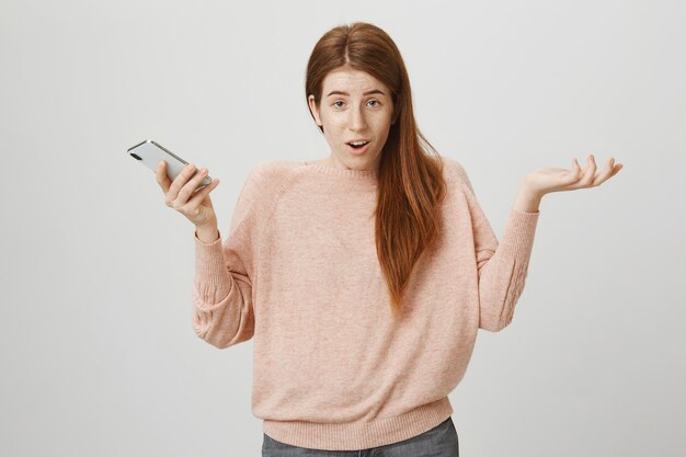 Mujer pelirroja despistada encogiéndose de hombros con teléfono en mano