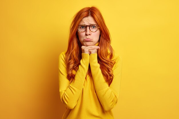 Mujer pelirroja disgustada y sombría, el labio inferior mantiene las manos debajo de la barbilla, expresa las emociones negativas que se siente ofendida, usa un jersey de manga larga.