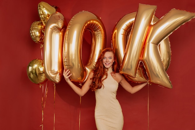 Mujer pelirroja en elegante vestido posando emocionada sosteniendo globos dorados en rojo