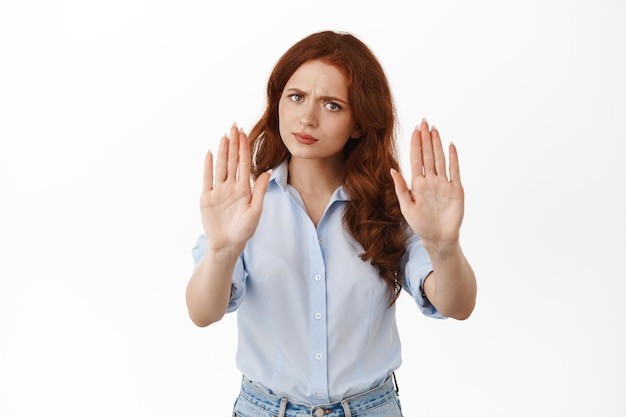 Para. La mujer pelirroja enojada extiende las manos y frunce el ceño, aléjate. Dije que no, rechazando algo, bloqueando una mala oferta, rechazando, de pie en blusa contra fondo blanco.