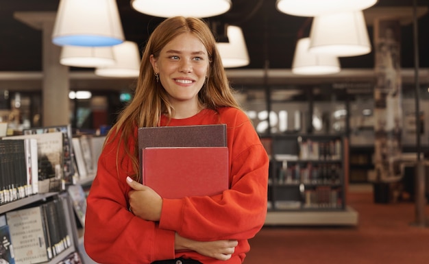 mujer pelirroja estudiando, sosteniendo libros en la librería y sonriendo.