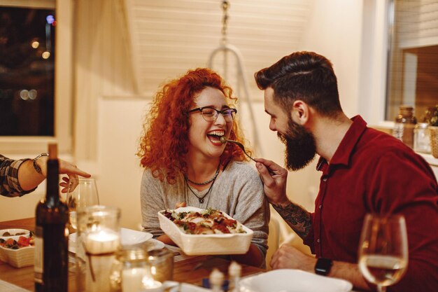 Mujer pelirroja feliz divirtiéndose mientras su novio la alimenta en la mesa del comedor