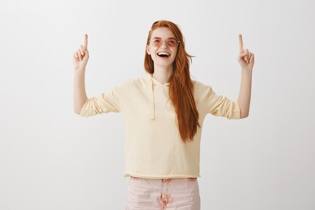 Mujer pelirroja feliz emocionada en gafas de sol apuntando con el dedo hacia arriba alegre