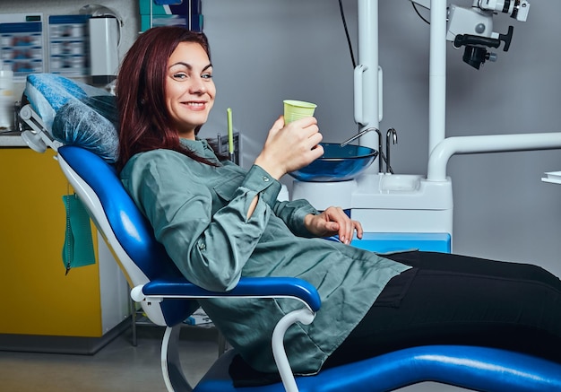 Mujer pelirroja feliz sentada en una silla de dentista sosteniendo una taza con enjuague bucal en una clínica.
