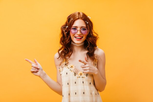 Mujer pelirroja con gafas de sol señala con el dedo a la izquierda. Mujer en top amarillo sonriendo sobre fondo naranja aislado.