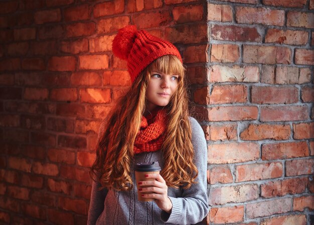 Mujer pelirroja con un gorro rojo y guantes posando sobre la pared de un ladrillo.