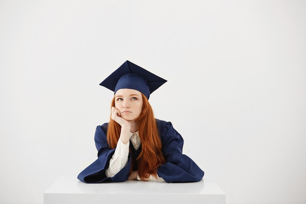 Mujer pelirroja graduada en manto pensando sentado sobre una superficie blanca