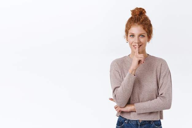 Mujer pelirroja hermosa y asertiva con el pelo rizado en un moño que usa un suéter acogedor que muestra el signo de silencio y una sonrisa tortuosa tiene una sorpresa para el novio que le pide a un amigo que guarde silencio, por favor