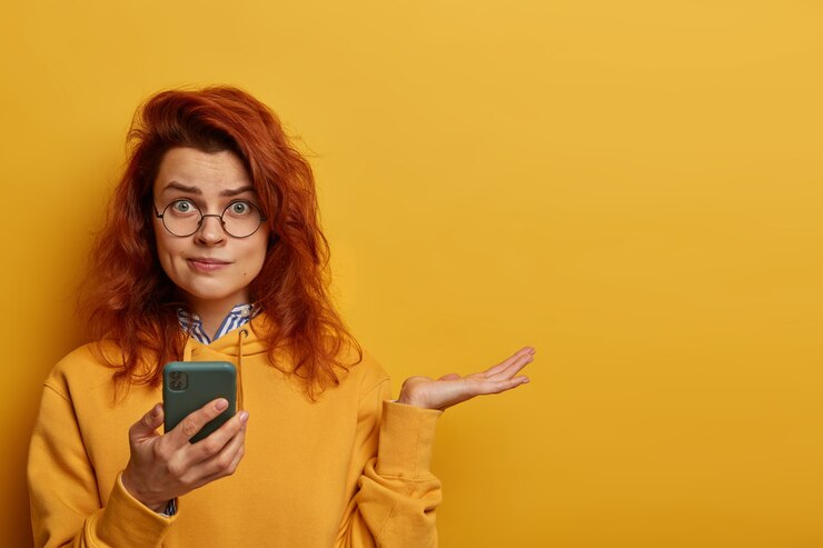Foto gratuito la mujer pelirroja indignada y desconcertada levanta la palma, piensa qué responder en el mensaje recibido, sostiene el teléfono móvil, usa gafas redondas y una sudadera con capucha, modela sobre una pared amarilla con un espacio en blanco a la derecha.