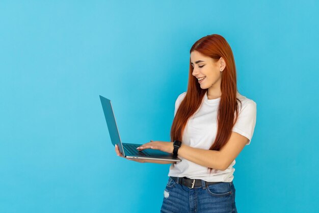 Mujer pelirroja joven emocionada mantenga la computadora portátil con la mano en la mejilla posando aislada en la pared azul turquesa