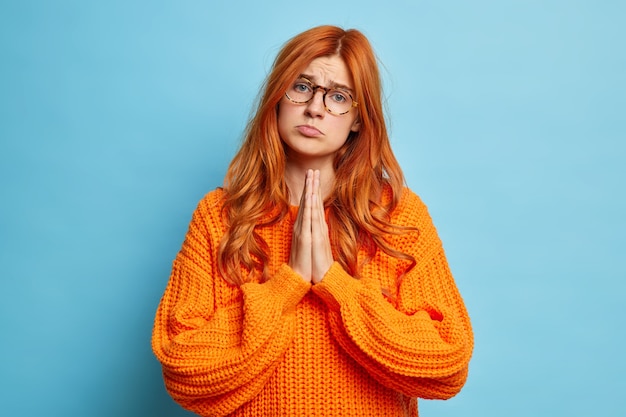 La mujer pelirroja lamentable frunce los labios y mantiene las manos en oración pide un favor tiene una expresión de descontento vestida con un jersey naranja casual.