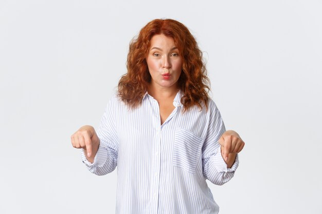 Mujer pelirroja de mediana edad tonta y linda que hace pucheros y da una pista señalando con el dedo hacia abajo, mostrando un banner con una oferta especial, haciendo un anuncio, de pie con un fondo blanco.