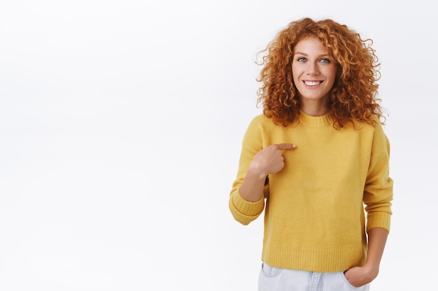 Mujer pelirroja con pelo rizado en suéter amarillo, apuntando a sí misma y sonriendo