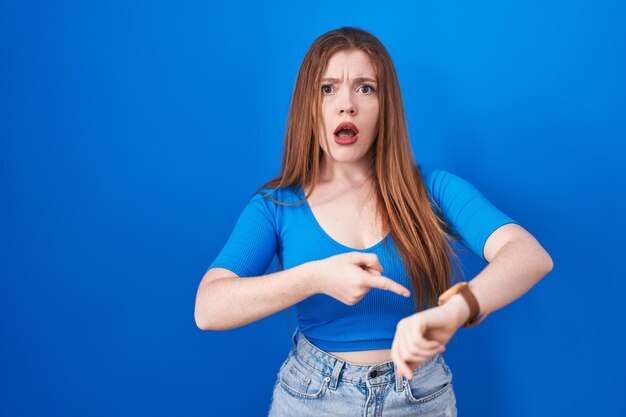 Mujer pelirroja de pie sobre fondo azul con prisa señalando el tiempo de reloj, impaciencia, disgustada y enojada por el retraso de la fecha límite