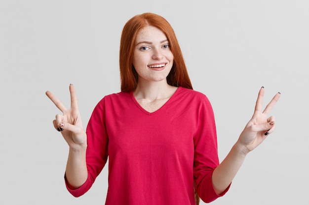 La mujer pelirroja positiva se divierte en el interior, hace gestos y muestra un signo bien, usa ropa casual roja, aislada sobre la pared blanca. Linda mujer con mirada positiva expresa su aprobación y felicidad