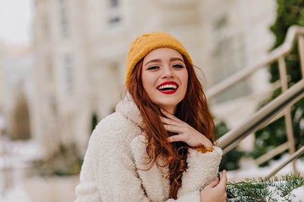 Mujer pelirroja positiva riendo en naturaleza borrosa. Chica de jengibre refinada sonriendo durante la sesión de fotos de invierno.