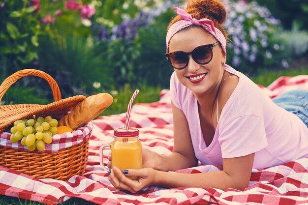 Mujer pelirroja positiva sentada en un banco con una cesta de picnic llena de frutas, pan y vino.