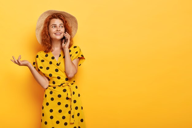 Mujer pelirroja sincera posando en vestido de lunares amarillo y sombrero de paja