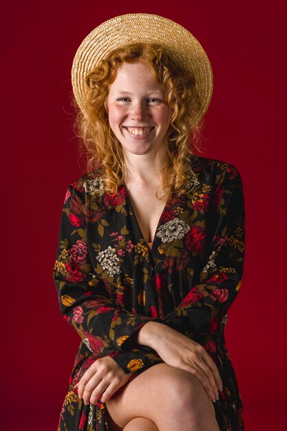 Mujer pelirroja con sombrero sonriendo y sentada con las piernas cruzadas