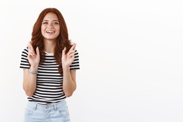 Mujer pelirroja soñadora linda ambiciosa en camiseta rayada que tiene fe, con suerte mirar la cámara, sonriendo anticipando el deseo hecho realidad, cruzar los dedos buena suerte, rezando fortuna, pared blanca