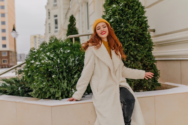 Mujer pelirroja soñadora posando en la calle en día de invierno. Foto al aire libre de feliz niña caucásica expresando emociones positivas.