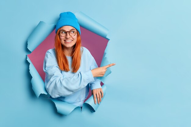 La mujer pelirroja sonriente complacida señala con el dedo en el espacio de la copia muestra una oferta especial o una venta de compras recomienda un buen descuento vestida con un elegante atuendo azul tiene un estado de ánimo feliz que se rompe a través del agujero del papel