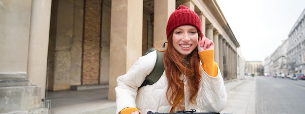 Foto gratuita una mujer pelirroja sonriente parada cerca del edificio alquila una escooter para viajar por la ciudad mientras está de turismo