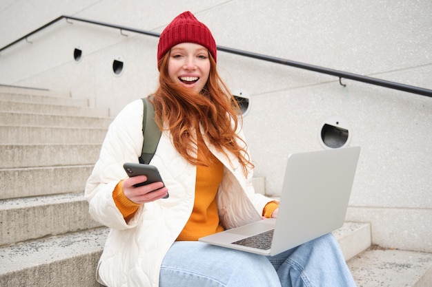 Foto gratuita mujer pelirroja sonriente con teléfono móvil y computadora portátil sentada en las escaleras fuera del edificio se conecta a pu