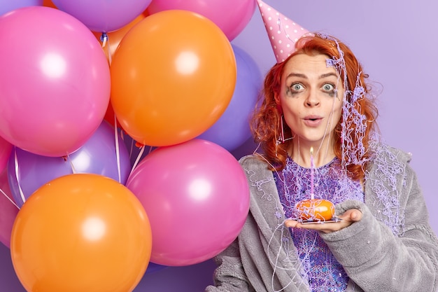 Foto gratuita mujer pelirroja sorprendida con maquillaje estropeado después de celebrar el aniversario mira sorprendida al frente sostiene cupcake y coloridos globos inflados aislados sobre pared púrpura