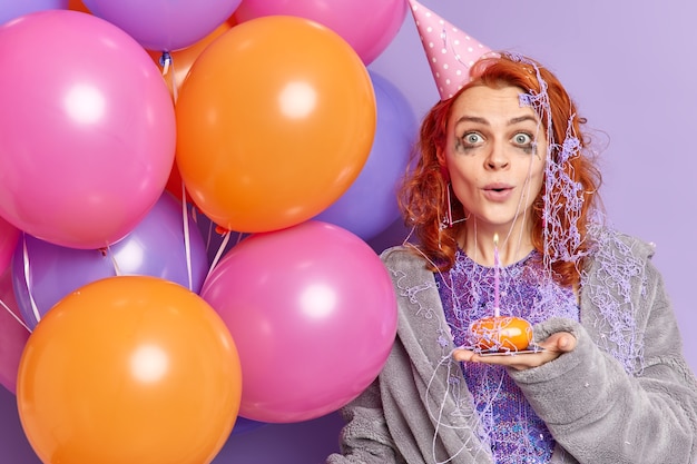 Mujer pelirroja sorprendida con maquillaje estropeado después de celebrar el aniversario mira sorprendida al frente sostiene cupcake y coloridos globos inflados aislados sobre pared púrpura