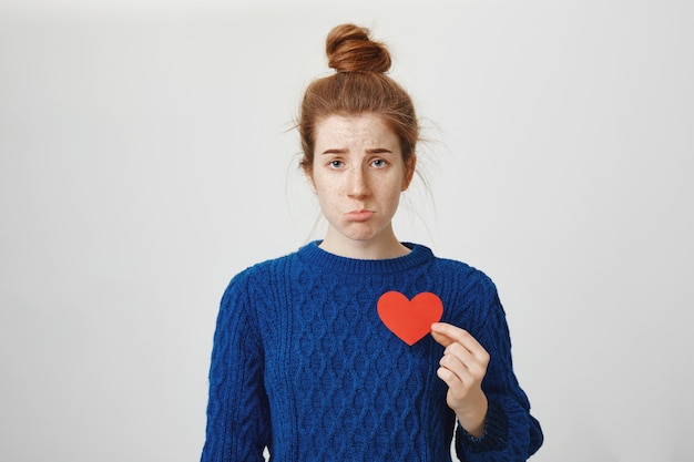 Mujer pelirroja triste con el corazón roto haciendo pucheros molesto, con cartel de corazón