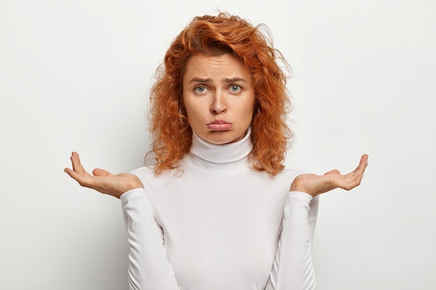 Foto gratuita mujer pelirroja triste y sombría frunce el labio inferior, toma una decisión seria, siente dudas e incertidumbre, extiende las palmas hacia los lados, vestida con un jersey blanco informal, sin estar segura de cómo resolver su problema