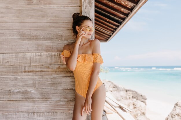 Mujer de pelo castaño de ensueño con gafas de sol descansando en el resort y disfrutando de vistas al cielo. Tiro al aire libre de hermosa chica delgada en traje de baño posando junto a la pared de madera con expresión de cara feliz.