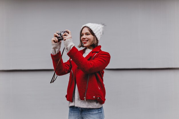 Mujer de pelo corto con gorro de punto y abrigo rojo toma fotografía en cámara retro.