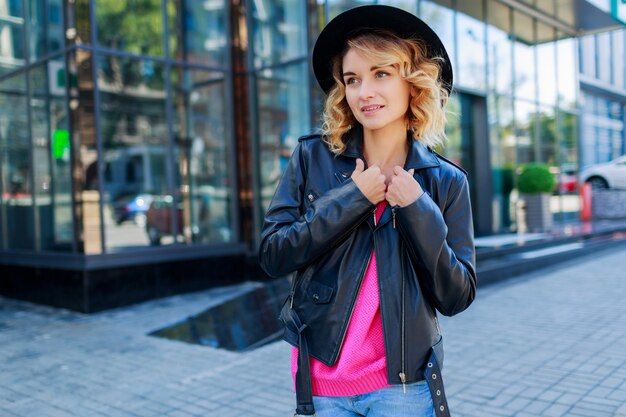 Mujer de pelo corto rubia pensativa caminando por las calles de la gran ciudad moderna. Traje urbano de moda. Gafas de sol rosa inusuales.