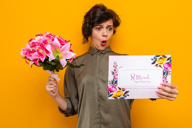 Mujer con pelo corto sosteniendo una tarjeta de felicitación y un ramo de flores mirando la tarjeta confundida y sorprendida celebrando el día internacional de la mujer el 8 de marzo
