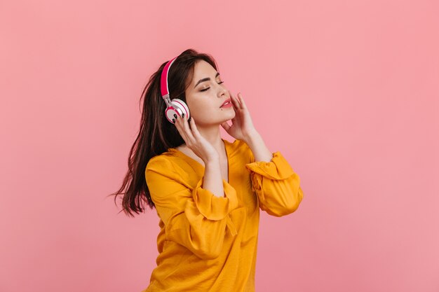 Mujer de pelo largo en blusa brillante y auriculares blancos y rosas escuchando música en la pared aislada.