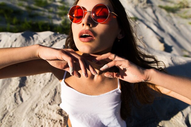 Mujer con el pelo largo en gafas de sol rojas se sienta en la arena blanca