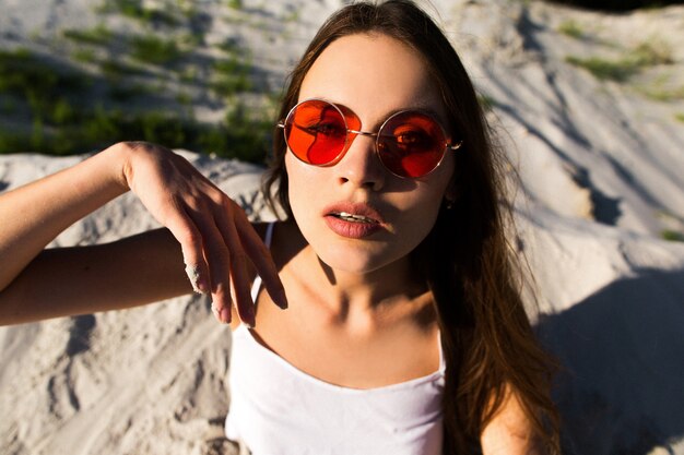 Mujer con el pelo largo en gafas de sol rojas se sienta en la arena blanca