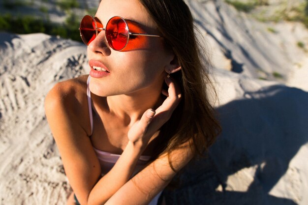 Foto gratuita mujer con el pelo largo en gafas de sol rojas se sienta en la arena blanca